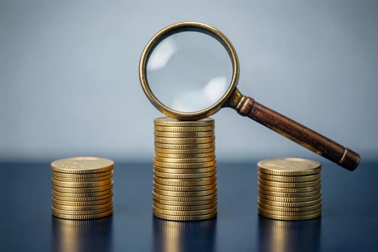 Três pilhas de moedas de ouro de tamanhos diferentes, com uma lupa focada na pilha maior.