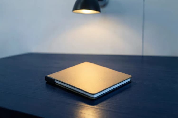 Um bloco de notas fechado sobre uma mesa de madeira escura, em plano médio, com luz de abajur vinda de cima.