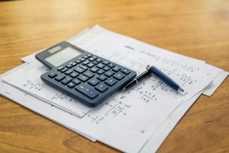 Um close-up de uma calculadora e uma caneta sobre papéis com números e símbolos, em uma mesa de madeira.